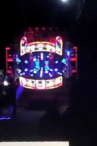 The video shows a large sound system with flashing lights at night. Silhouetted figures are visible in the foreground and on the sides. The sound system is mounted on a vehicle, likely a truck, and is decorated with colorful lights. The lights are primarily red, blue, and yellow, and they flash in a rhythmic pattern. The overall atmosphere is one of a nighttime event or party.