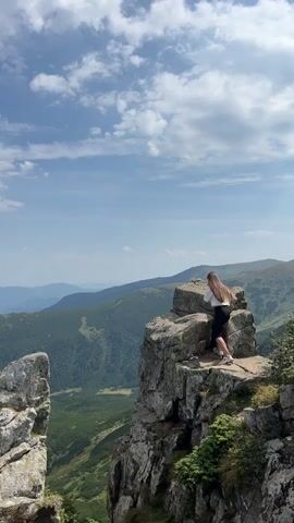 A young woman with long brown hair is climbing on a rocky outcrop. She is wearing a light-colored t-shirt and dark shorts. The background is a mountainous landscape with green valleys and a blue sky with white clouds. The woman is actively moving, adjusting her position on the rocks.