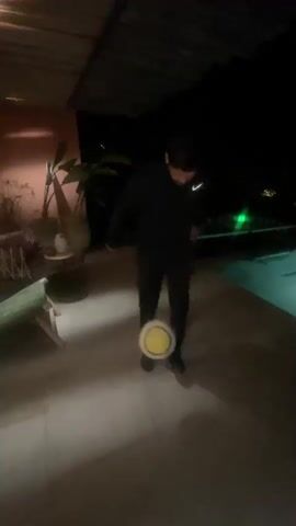 A man wearing black clothing juggles a soccer ball near a pool at night. The man uses his feet and knees to keep the ball in the air. The area is dimly lit, with some greenery and a covered porch visible. The pool is illuminated with a green light.