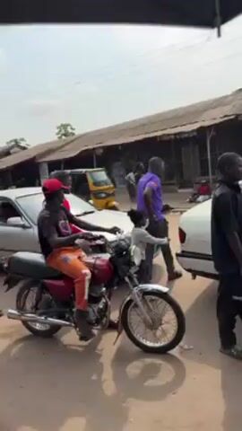 The video shows a busy street scene with several people, cars, a motorcycle, and a hand-pulled cart. A man is sitting on a red motorcycle with orange pants and a red cap, seemingly waiting. A man dressed in black is pulling a cart filled with various small items. There are two white cars parked on the street. A yellow three-wheeled vehicle is partially visible in the background. Other people are walking around, including one in a red jacket and another in a purple shirt.  A black goat wanders into the scene. The background shows buildings with corrugated metal roofs, and some people are sitting on the ground near the buildings. The street appears to be unpaved. The sky is overcast.