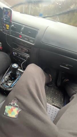 The interior of a car, likely a Volkswagen, is shown from the passenger seat point of view. The driver is wearing black sweatpants with a patch and black sneakers. The car is driving on a wet road. The dashboard is visible, including the steering wheel, radio, and a phone mounted on the dashboard. An air freshener hangs from the rearview mirror. The weather appears overcast. The car is moving, based on the changing scenery through the windshield. Music is playing inside the car.