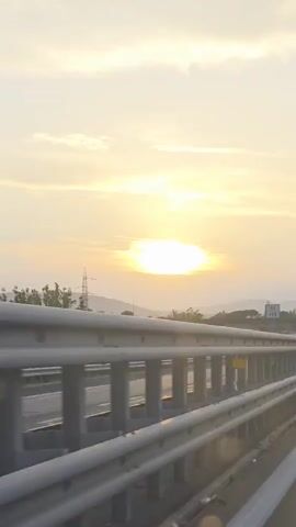 The video shows a view from a moving vehicle on a highway at sunset. The sun is a bright, large orb in the sky, casting a warm glow and creating lens flares. The sky is filled with soft, diffused clouds. In the foreground, a metal guardrail runs along the side of the highway. The highway itself is visible, with other vehicles, including cars and a camper van, driving in the same direction. In the distance, hills and power lines can be seen against the horizon. The overall atmosphere is serene and captures the feeling of a road trip during golden hour.