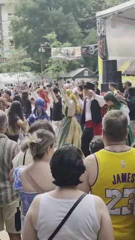 A crowded outdoor event is taking place, possibly a festival or fair. In the foreground, several people are visible from behind, facing away from the camera. They appear to be spectators, with one person wearing a yellow Lakers basketball jersey. Behind them, a group of people are dancing in a circle, holding hands. Some of the dancers are wearing traditional or folk costumes, including a woman in a long, flowing dress and headscarf, and a man in a vest and hat. Others are dressed in modern clothing. In the background, there are tents, trees, and a building, as well as a stage with speakers. The overall atmosphere appears lively and festive.