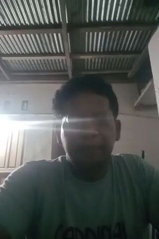 The video is a low-resolution, shaky selfie of a man. He is wearing a light blue t-shirt and appears to be indoors, as evidenced by the corrugated metal ceiling visible above him and the wall and door frame in the background. A light source, possibly a window or lamp, is casting a strong glare, obscuring the man's face in the first few frames. His facial features are more discernible in the later frames, revealing a man of middle age with dark hair and a mustache. The man is looking directly at the camera and speaking, though his voice is not transcribed here. The video cuts out abruptly.