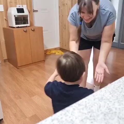 A woman and a young child are in a room with wood-paneled walls and a wood-effect laminate floor. The woman is wearing a blue t-shirt and black shorts, and white socks. The child is wearing a dark t-shirt. The woman is bent over, holding out her hands to the child. The child is facing the woman, also with hands outstretched. On a wooden cabinet to the left, there is a white air fryer. In the background, there is a white door and more wood paneling.