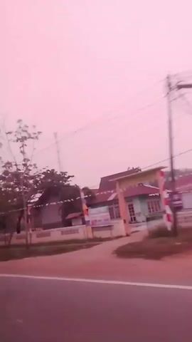 The video is a first-person perspective shot from a moving vehicle, showing a roadside scene. The environment appears to be a rural or suburban area with houses, trees, and utility poles visible. The sky is a soft pink, suggesting either dawn or dusk, or possibly a filter effect. Several houses with distinct architectural styles and tiled roofs are present. A white truck and a turquoise truck pass by in the opposite direction, indicating traffic on the road. The footage is slightly blurry due to the movement of the camera and the passing vehicles. There are no people clearly visible in the video. The road surface is a dark gray asphalt. The overall atmosphere is calm and transient, capturing a slice of everyday life on a road.