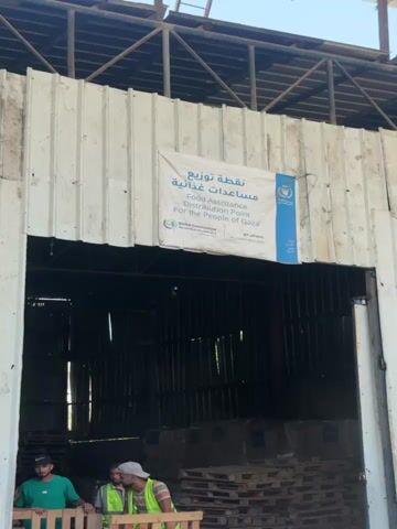The video shows a line of people in front of a large warehouse-like building. The building has corrugated metal walls and an open entrance. A banner above the entrance reads "World Assistance Distribution Point For the People of Gaza" in English and Arabic. There are stacks of boxes wrapped in plastic on wooden pallets. Several people are standing in line, and some are interacting with individuals who appear to be distributing goods. The overall scene suggests a humanitarian aid distribution point.