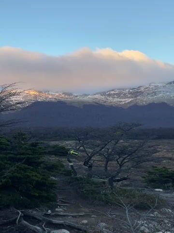 The video shows a mountainous landscape. The foreground consists of a grassy area with scattered trees and shrubs. Some of the trees are bare, while others are evergreen. In the middle ground, there's a small stream flowing through the landscape. The background is dominated by a range of mountains with snow-capped peaks. Some of the peaks are partially obscured by clouds. The sky is blue with a few scattered clouds. The overall tone of the video is serene and peaceful.