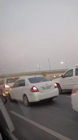 A traffic jam on a highway is depicted from inside a vehicle. White cars, including SUVs and sedans, are stationary on a multi-lane road. A pedestrian overpass is visible in the background, along with streetlights. The sky is overcast and hazy. The video is filmed from a side-facing perspective, capturing the movement of vehicles as the filming vehicle inches forward.