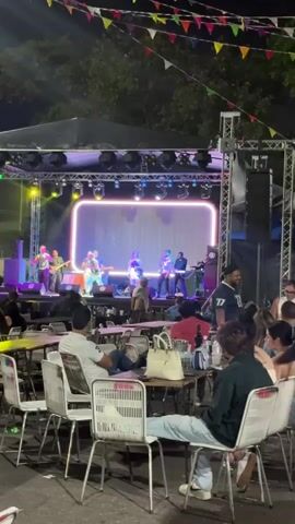 The video shows a live band performing on an outdoor stage at night. The band, named "LA SUAVECITA BANDA" is illuminated by stage lighting, and a large screen behind them displays the band's name. The audience sits at tables in front of the stage, some enjoying drinks while watching the performance. The area is decorated with colorful pennant banners strung above the tables. Servers are moving among the audience, attending to patrons.