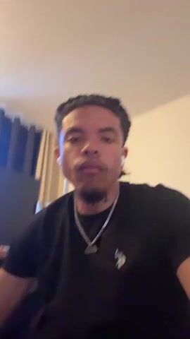 A young man with braided hair and tattoos on his neck is wearing a black t-shirt and a silver chain. He is sitting in a room with curtains in the background. He is looking at the camera and makes various facial expressions. In one shot, he runs his hand through his hair. In another shot, he touches his neck. The man is also wearing white earbuds.
