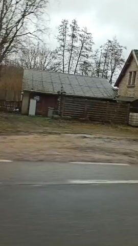 The video shows a view from a moving vehicle, likely a car, driving along a road in a rural area. The buildings on either side of the road are generally old and made of stone or wood. The weather appears to be overcast and possibly wet, as indicated by puddles on the road. The video passes by various structures including a building with a colorful graffiti mural, another building with a closed shutter door, a business with stacked containers, and a two-story house. Bare trees are visible against the cloudy sky.