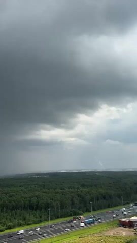The video shows a wide-angle view of a landscape under a stormy sky. The foreground features a multi-lane highway with several vehicles traveling on it. Beyond the highway, there is a dense forest. In the distance, a visible rain shower is occurring, indicated by a grey curtain of rain falling from the dark, heavy clouds. The sky is overcast with thick, dark clouds, suggesting impending or ongoing bad weather. There are some buildings visible in the far distance, partially obscured by the rain. The overall mood is dramatic and indicative of a stormy day.