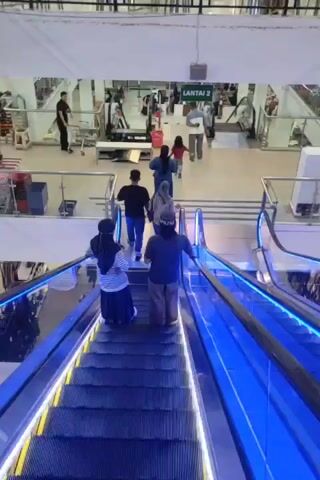 The video begins with a view looking down an escalator with blue lights. There are two people on the escalator in front of the camera, one wearing a hijab and the other wearing a head covering and a long-sleeved shirt. They are facing away from the camera. In the background, several other people are visible on the level above, walking towards another escalator and stairs. One person is pushing a shopping cart. A sign above the entrance to the upper level reads "LANTAI 2." The environment appears to be a shopping mall or department store, with displays and merchandise visible in the background. The escalator is moving downwards. The lighting is bright and the scene is well-lit.