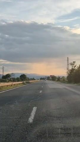 The video shows a view from a vehicle traveling on a highway during daylight. The sky is partly cloudy with a sunset glow visible in the distance. There are power lines and utility poles alongside the road, as well as trees and vegetation. The road itself appears to be asphalt with white lane markings and some visible wear and tear. There are a few other vehicles on the highway in the distance.