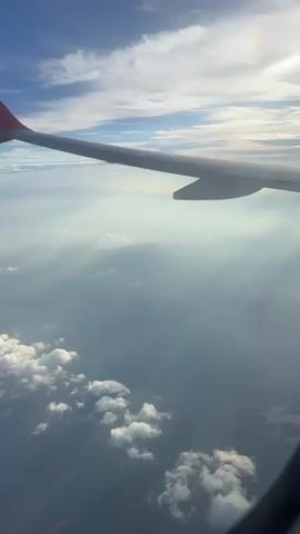 The video captures an aerial view from an airplane window. The wing of the airplane is visible in the foreground, extending from the left side of the frame towards the right. The wing has a red tip. Below the wing, a vast expanse of clouds is seen, with some cumulus clouds scattered closer to the airplane and a more hazy layer of clouds and mist stretching towards the horizon. The sky above is blue with wispy white clouds. The sunlight is filtering through the clouds, creating rays of light and casting a soft glow over the landscape below. The overall impression is one of peaceful flight over a cloudy sky.
