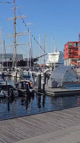 The video shows a harbor with several boats docked. A large, multi-masted sailing ship is prominently featured on the left side of the frame. Several smaller sailboats and a ferry are also visible. In the foreground, a wooden walkway leads to a pier with a clear, dome-shaped structure. Modern, red buildings with visible windows are situated on the right side of the harbor. The sky is clear and blue, suggesting a sunny day. The water is calm, reflecting the boats and structures.