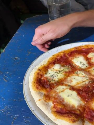 The video opens with a close-up of a margherita pizza on a white plate, resting on a textured blue table. The pizza has a golden-brown crust, red tomato sauce, melted white mozzarella cheese, and is sprinkled with dried herbs, garnished with two fresh green basil leaves. A person's right hand with visible knuckles is resting on the blue table to the left of the pizza. In the background, a clear glass is partially visible. The camera then moves to show a man's torso dressed in a black t-shirt, before panning up to his face. The man has short light brown hair and is wearing dark-rimmed glasses, looking directly at the camera with a subtle smile.