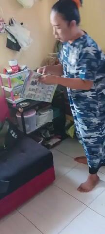 A person wearing a blue and white camouflage patterned dress stands in a room and holds up a booklet. The booklet has colorful images and text on its pages. The room is decorated with various items, including bags of snacks hanging from the wall and shelves filled with various products. A green chair is visible in the background.