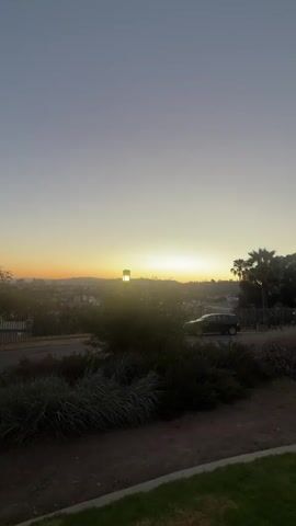 The video opens with a wide shot of a sunset over a city skyline. The sky transitions from a soft orange near the horizon to a pale blue at the top. In the foreground, there is a park with green grass, bushes, and trees. A car is visible driving on a road in the middle ground, and a street lamp stands tall to the left of the car. As the video progresses, the camera pans to the right, revealing several groups of people spread out on the grass, enjoying a picnic. They are sitting on blankets, some are eating, and others are talking. The overall atmosphere appears relaxed and communal, with the golden hues of the sunset casting a warm glow over the scene.