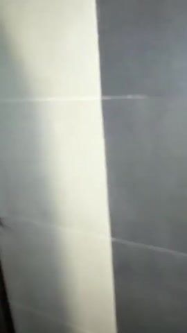 A spider is seen on a white tiled wall next to a dark brown door frame. The spider is dark brown and has eight legs. It is positioned on the wall, and its legs are spread out. The lighting in the video is dim, with a bright light casting a shadow on the wall from the left side of the frame. The spider is visible from the beginning of the video until the end.