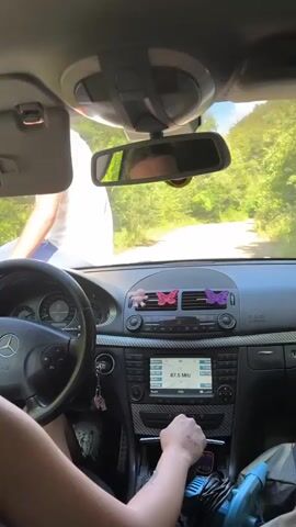 The video is shot from inside a car, looking out the windshield. A person is seen sitting in the driver's seat, their arm extended toward the gear shift. A second person, dressed in a white shirt, is standing outside the car on the driver's side, partially visible through the windshield and side window. The car's dashboard is visible, with a radio display showing "87.5 MHz". Outside the car, a dirt road winds through a wooded area with green trees. The sun is shining, casting shadows. The overall scene appears to be a brief moment during a car journey in a natural setting.