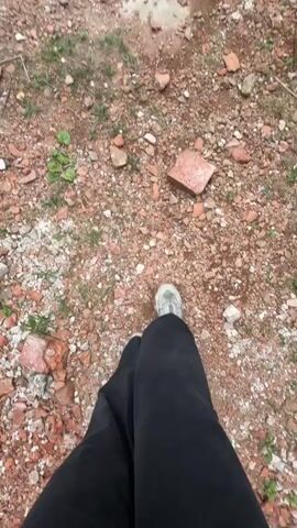 The video shows the inside of a ruined brick building, likely a church or similar structure. The camera starts at ground level, focusing on the ground littered with broken bricks and some patches of green vegetation. The camera then tilts upward to reveal the interior of the building, showcasing the crumbling brick walls and arches. The sky is visible through the collapsed roof. There is a person in the video, wearing black pants and light-colored shoes, walking on the brick-covered ground. The person's legs are visible in the foreground. The overall atmosphere suggests abandonment and decay.
