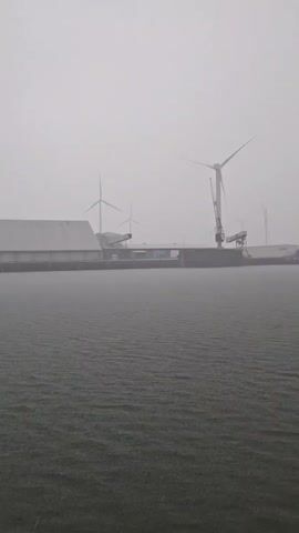 The video shows a rainy seascape, viewed from the perspective of someone on a vessel. In the distant background, several wind turbines are visible, along with what appears to be industrial buildings. As the camera pans slightly to the right, a large offshore platform comes into view, its support legs extending high into the misty sky. The rain is dense, creating a visual haze and causing visible ripples on the water's surface. The foreground includes parts of the vessel's structure, specifically white beams, framing the view of the landscape.