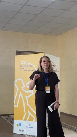 A woman stands in front of a yellow banner with white graphics and Ukrainian text. She is wearing a dark brown jumpsuit and a lanyard with a badge. The banner has text that appears to say "Миронівка: місто, яке говорить" which translates to "Myronivka: a city that speaks". There are also logos on the banner, including one that looks like "МХП-Громади". The woman is gesturing with her left hand and holding a small book or notepad in her right hand. She has blonde hair and appears to be speaking at an event or presentation. The background shows a wall with textured wallpaper and a ceiling with acoustic tiles. The lighting suggests it is daytime, and the indoor setting indicates an event space.