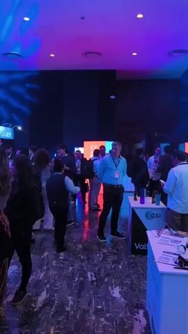 The video shows a crowded room, possibly at a conference or networking event. Numerous people are present, dressed in business casual or professional attire, and engaged in conversations. Some individuals are holding drinks. There are several display booths set up, with logos such as "Lulubit" and "GSI" visible. The lighting is a mix of blue and pink, creating a vibrant and somewhat formal atmosphere. The floor has a dark, marbled pattern.