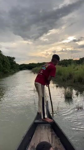 A young man wearing a red t-shirt and light-colored pants stands at the front of a narrow, dark-colored boat, poling it through a wide, calm river. The riverbanks are lined with lush green vegetation and trees, and the sky above is filled with dramatic, overcast clouds with patches of sunlight breaking through. Another person, partially visible, sits in the boat behind the young man. The young man uses a long wooden pole to propel the boat forward, dipping it into the water and pushing off the riverbed. He appears focused on his task, and the boat glides smoothly through the water. The overall scene is one of peaceful travel through a natural, scenic environment.