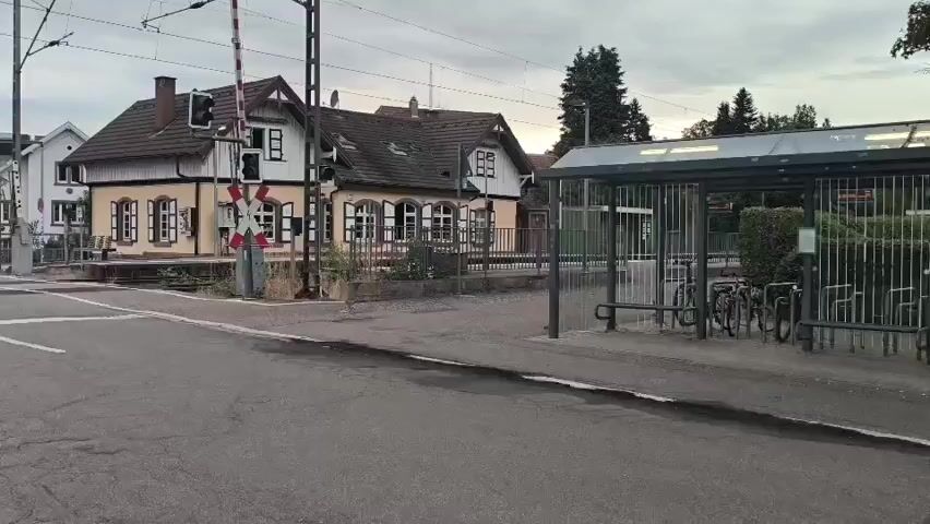 The video shows a railway crossing with a gate. A yellow and white building is visible behind the gate. A bus stop shelter with bicycles parked inside is on the right side of the frame. The sky is overcast.