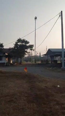 The video pans across an outdoor scene with dry, brown grass in the foreground. In the middle ground, there is a dirt path and several structures that appear to be small shops or food stalls, some with open fronts and seating areas. A tall pole with a speaker or siren is visible in the distance, along with power lines. Further back, there are trees and some residential-looking buildings. The sky is clear and pale, suggesting either early morning or late afternoon light. The overall impression is a somewhat unkempt, rural or semi-rural environment, possibly a market or gathering place. There are no visible people in the immediate foreground, but two figures can be seen walking in the distance on the path. A small red and yellow flag is planted in the ground in the foreground.