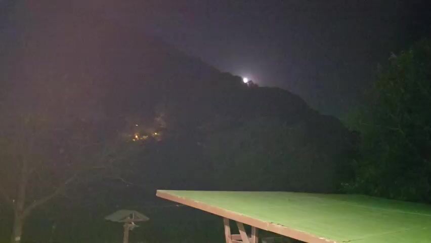 A night video shows a hill with a bright moon above it. There are some lights on the hill and some trees in the foreground. The video is shaky and unclear due to low light conditions.
