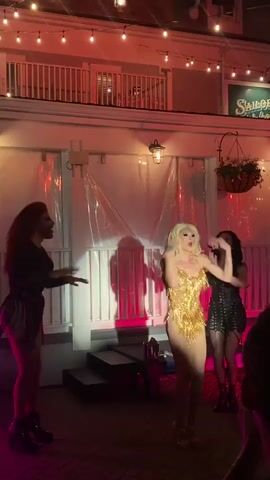 The video shows three drag queens performing on an outdoor stage at night. The stage is illuminated by pink and red lights, and there are string lights overhead. In the background, there is a building with a balcony and a sign that reads "Sailor's Rest." The drag queens are all dressed in elaborate costumes and are dancing and posing for the camera. The queen in the center is wearing a gold sequined leotard with a fringe skirt. The queen on the left is wearing a black outfit with fringe detailing. The queen on the right is wearing a black corset and skirt. The audience is visible in the foreground, with some people holding up phones to record the performance.