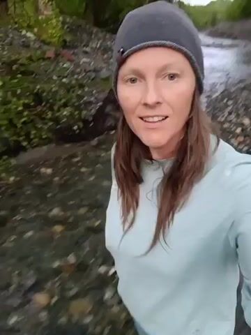 The video shows a smiling woman taking a selfie video in a rocky riverbed. She wears a gray beanie, a light blue long-sleeved shirt, and blue pants. Her reddish-brown hair hangs down to her shoulders. The river flows behind her, and the banks are covered in rocks and some green vegetation. The lighting is somewhat dim, possibly indicating it is early morning or late evening. The video appears to be hand-held, and slightly shaky.