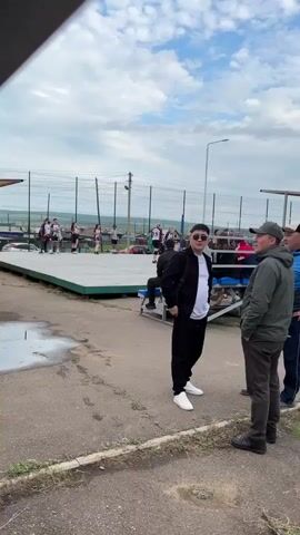 The video shows an outdoor scene, likely a sporting event or gathering. In the foreground, three men are engaged in conversation. The man on the left wears a black jacket and pants, a white t-shirt, and white sneakers. The man in the middle is in a green jacket, dark pants, and a baseball cap. The man on the right wears a blue jacket and dark pants. Behind them, a raised platform is visible, with people on it. Further back, there are bleachers with spectators. In the background, a volleyball game appears to be in progress, with players on the court and more people in the stands. The overall environment is outdoors with a cloudy sky. The video also shows parts of a large metal structure, possibly part of the stadium's infrastructure, and a grassy area beyond the paved ground. A young girl in a black hoodie is also visible in the frame, facing away from the camera.