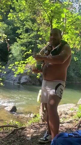 A man stands shirtless by a river, wearing white shorts. He holds a can in his left hand and a slice of food in his right, bringing it to his mouth to eat. He wears sneakers. A towel or fabric is draped over his shoulders. Trees and rocks surround the river.