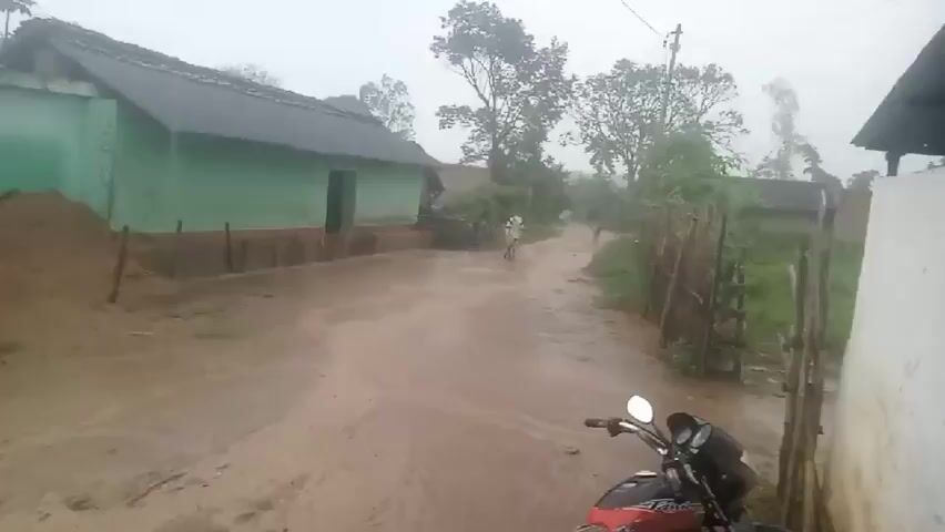 The video shows a rural area during a heavy rainstorm. The ground is muddy and flooded, with water flowing down a dirt road. A green building with a dark roof is visible on the left, with a large pile of sand next to it. Trees and vegetation are present in the background. A motorcycle is parked on the right side of the road, partially submerged in water. The overall scene depicts a flooded rural environment due to heavy rainfall.