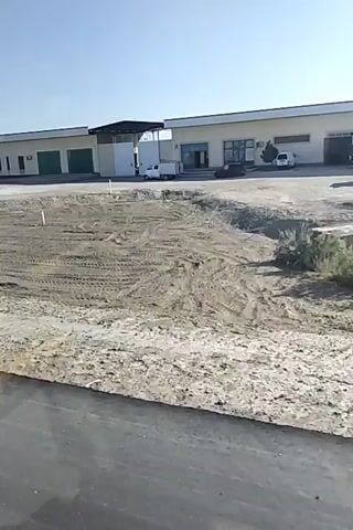 The video shows a construction site with several buildings in the background. The foreground consists of a large area of dirt with tire tracks, indicating recent activity. One building appears to be a warehouse with large green doors. Another building is a long, low structure with multiple windows. The sky is clear and blue. There are a few vehicles visible in the background, including a white van and a dark-colored car. The overall impression is of a developing industrial or commercial area.
