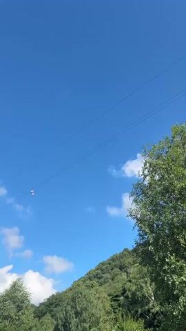 A person is zip-lining across a clear blue sky with a few scattered clouds. The zip-liner appears to be wearing a red and white top and dark bottoms. The zip-line is supported by two parallel wires. A lush green, tree-covered hillside is visible in the lower right portion of the frame. The camera is angled upwards, capturing the person in mid-air.