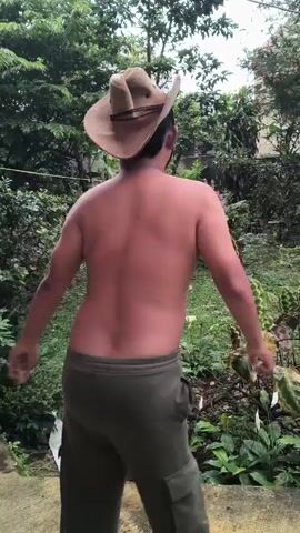 A shirtless man wearing a cowboy hat and green pants is dancing in a backyard. He is facing away from the camera but turns his head to smile at the end of the video. The backyard is overgrown with plants and trees.
