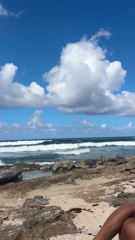 The video shows a man surfing in a choppy ocean. The surfer is wearing dark swim trunks and is riding a wave towards the rocky beach. The sky is blue with puffy white clouds. The waves are white-capped and moderately sized. The beach is sandy and rocky with some sparse vegetation. The camera is positioned at a low angle, looking out over the beach towards the ocean. The video captures the movement of the surfer and the waves, as well as the scenic beauty of the beach and sky. A leg is visible in the foreground, presumably belonging to the videographer.