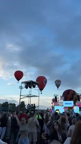 The video shows an outdoor festival or concert with a crowd of people gathered. In the background, there are several hot air balloons in the sky. One red balloon with a logo is prominent, alongside another red balloon and a multi-colored balloon. A stage or scaffolding structure with lights and equipment is visible, with some individuals working on it. The crowd appears to be enjoying themselves, with many people raising their hands and taking photos or videos. The sky is partly cloudy, suggesting it is either early morning or late afternoon.