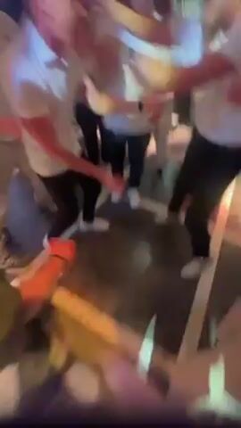A group of men in white shirts are huddled together, seemingly dancing and cheering in an indoor setting with colorful lighting. They are holding each other’s hands and arms, and appear excited or celebratory. The background is blurry but indicates a bar or club atmosphere.