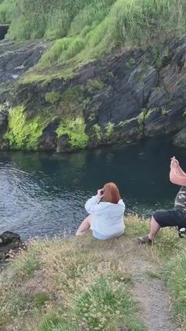 A woman with red hair wearing a white jacket and denim shorts sits on a grassy cliff overlooking a body of water. She is holding her hands up near her face. To her right, a man wearing a camouflage tank top and dark shorts is also sitting and holding his hands up, possibly taking a photo or video. The cliff is covered in grass and rocks with green moss. The water is dark blue and calm. The scene is outdoors, likely during daylight.