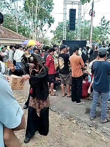 The video shows a street scene with a crowd of people. In the foreground, a person in a black costume and a mask resembling a demon or mythical creature is holding a cardboard box with the "Vit" logo. The masked person is gesturing towards the camera. Behind them, a large crowd of onlookers is gathered, with some standing and others sitting. There is a stage with speakers set up in the background, suggesting an outdoor event or performance. The environment appears to be a village or rural area with houses and trees visible. The overall atmosphere seems to be that of a lively gathering or festival.
