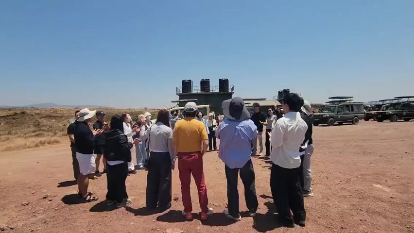 A group of about 15 people stand in a circle on a red dirt field on a sunny day with a clear blue sky. They appear to be listening to someone, perhaps a tour guide, as they are facing toward the center of the circle. Some people are wearing hats or caps for sun protection. In the background is a building with a vehicle parked next to it. To the right, several safari vehicles are parked. Some of the group are clapping their hands.
