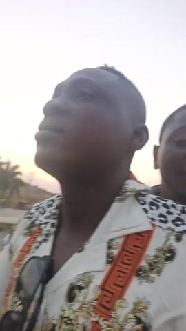 The video features two young men. The man in the foreground, with short hair, is wearing a patterned white shirt with sunglasses clipped to the front. He appears to be looking upwards. Behind him, another young man smiles slightly, mostly out of focus. The background includes trees and a pale sky, suggesting it might be late afternoon or early morning. Strong wind noise is present, indicating an outdoor setting with significant wind.