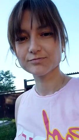 This is a short video of a young woman standing outside. She is wearing a pink tank top with yellow and white text on it, the text is partially obscured but appears to say "Baby". She has brown hair with bangs and is wearing hoop earrings and a thin necklace. The woman is looking at the camera and has a neutral expression.

The setting appears to be a rural or suburban area. In the background, there is a fence, a building that looks like a house, and trees. The sky is clear and blue, suggesting it is daytime. The lighting indicates it is likely late afternoon or early evening, given the warm tones and shadows. There are no specific events or activities taking place, it seems to be a casual recording of the woman.